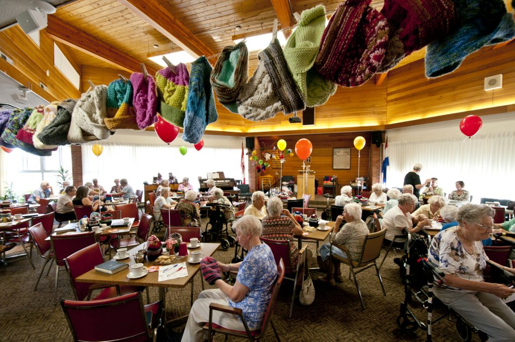 A photo inside of Harmony Hall during a festive knitting event for Christmas. There are knitted items hung on a string and many elderly women seated around the room.