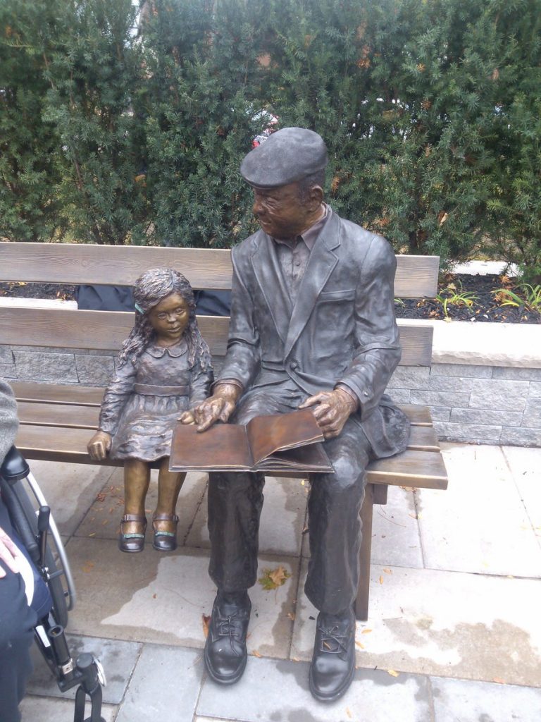 A photo of two bronze statues. A elderly man is reading a story to a young girl. They are both sitting on at the edge of a wooden bench.
