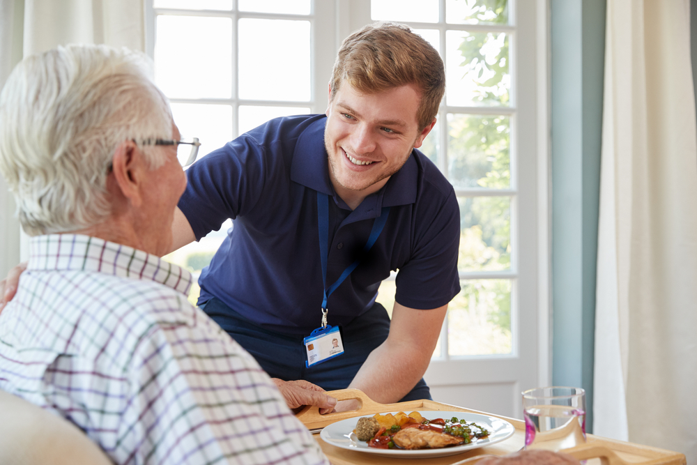 A photo of a male embracing their elderly relative in the Manors. The elderly man is also enjoying a nutritious meal while conversating with his relative.