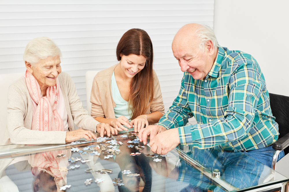 A photo of a elderly couple and their relative completing a puzzle in their apartment. They are happily piecing this puzzle together.