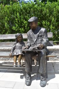 A picture of bronze statues; a older Dutch Man reading a book to a young girl bronze. Both are sitting on a bench in front of the main entrance.